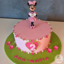 Tort roz cu verde vesel cu Șoricica Minnie - Minnie Mouse, îmbrăcată într-o rochiță elegantă, cu volănașe și buline albe, decorat cu șiraguri verzi cu multe, multe floricele roz și o plăcuță în formă de inimă