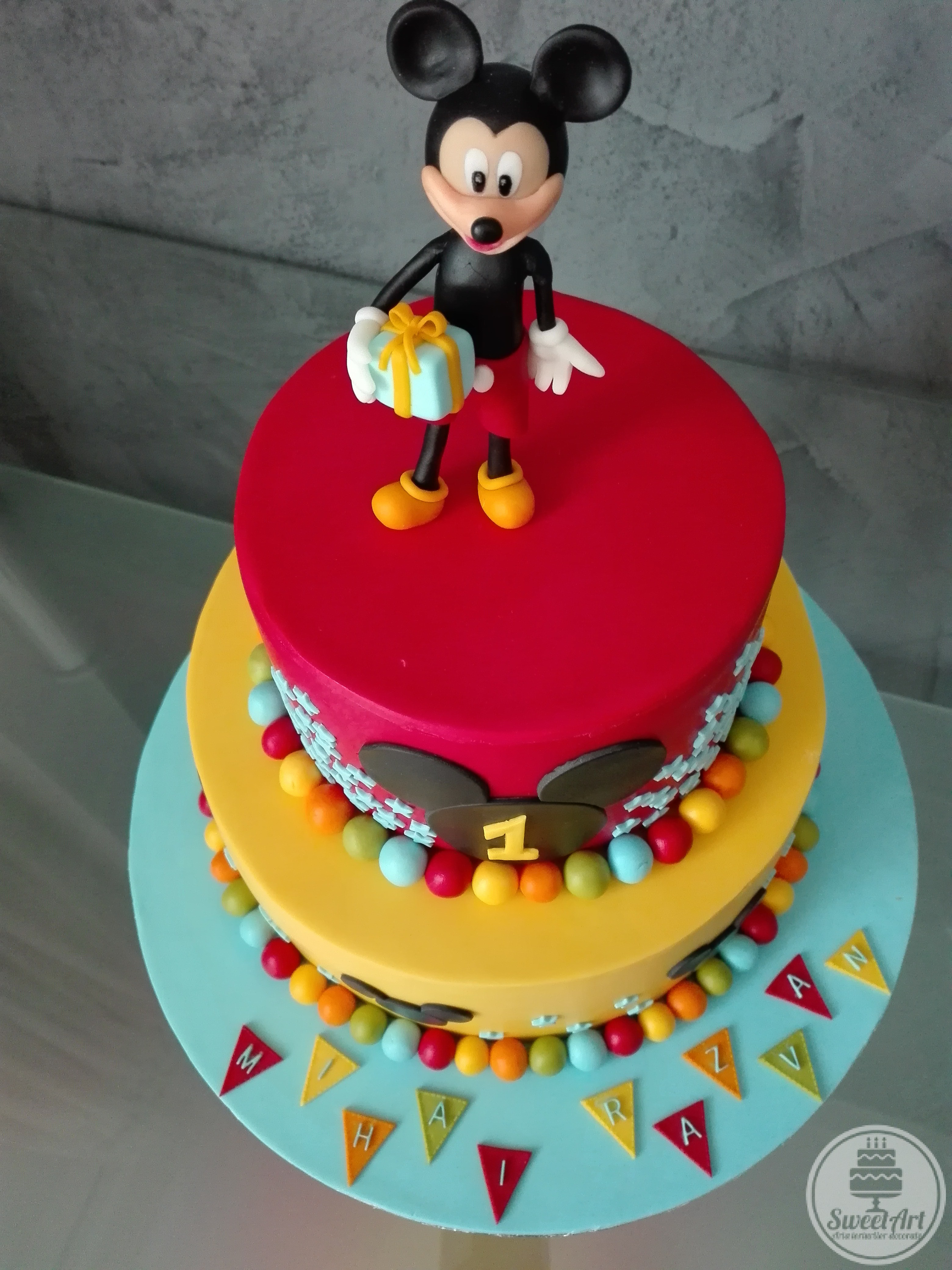 Tort cu Șoricelul Mickey - Mickey Mouse cu un cadou în mână, capul lui Mickey 2D și nunumărate steluțe bleu și bile colorate: roșii, bleu, verzi, portocalii și galbene