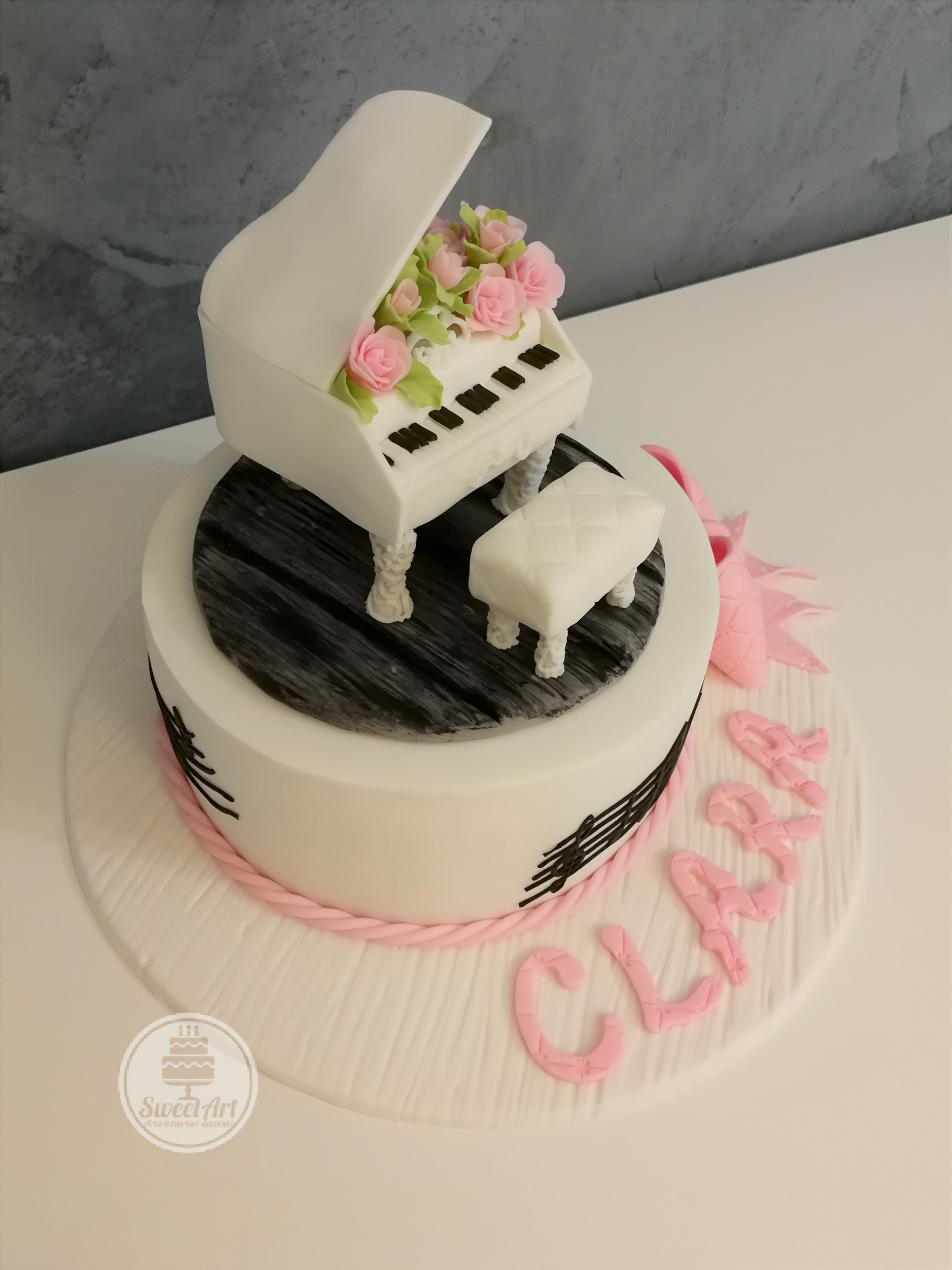 Tort pian cu aranjament cu trandafiri roz și frunze și banchetă albă pian pe parchet lemn negru, portativ cu note muzicale, fundă roz, tort alb pe platou lemn alb